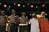 Five men are standing on stage, arm in arm; three are wearing traditional Rwandan attire with bare torsos, while the other two are covered in traditional Moroccan jellabas.