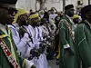A group of men wearing traditional white-and-green jellabas is walking in the Gnaoua parade, playing qraqeb and smiling.