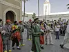 Several men are walking down a street in colorful traditional attire, playing the qraqeb, while onlookers on the side take pictures and wave to them.
