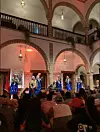 Several men are wearing blue jellabas, singing and dancing around an older man sitting among them, playing the guembri.