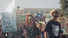 People at a street protest hold signs and walk forward in bright daylight.