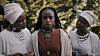 Marima Wanjiru as Anam stands between two women dressed in white headwraps and layered jewelry in a still from the Kenyan psychological thriller Anam’s Wake.