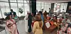 Guests standing and sitting in a bright lounge with large windows and colorful chairs.