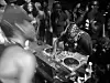 Crowd surrounds a DJ booth with turntables and mixers in a dark club setting.