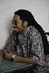 Mehdi Nassouli is wearing sunglasses and a batik grey and white shirt, sitting at a table with green tiles and smiling.