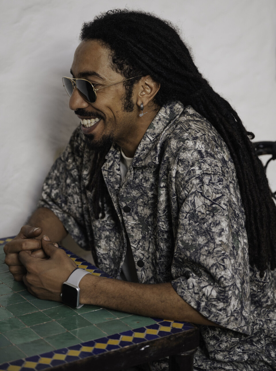 Mehdi Nassouli is wearing sunglasses and a batik grey and white shirt, sitting at a table with green tiles and smiling.