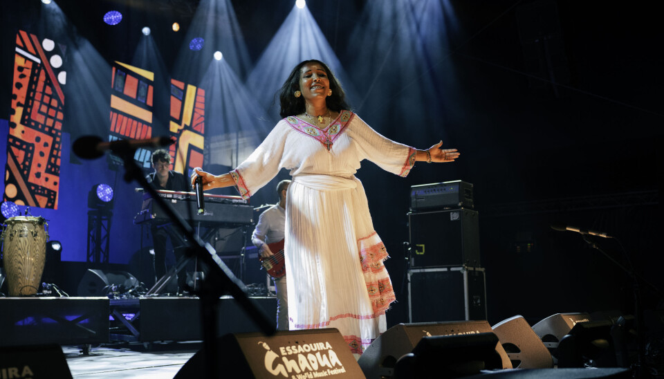 An Ethiopian woman wearing a white traditional dress is on stage, her arms spread out, white as she dances the traditional shoulder dance.