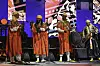 Four men are on stage in traditional red and green jellabas, two are playing the qraqeb, one the tleb drum, and one the guembri.