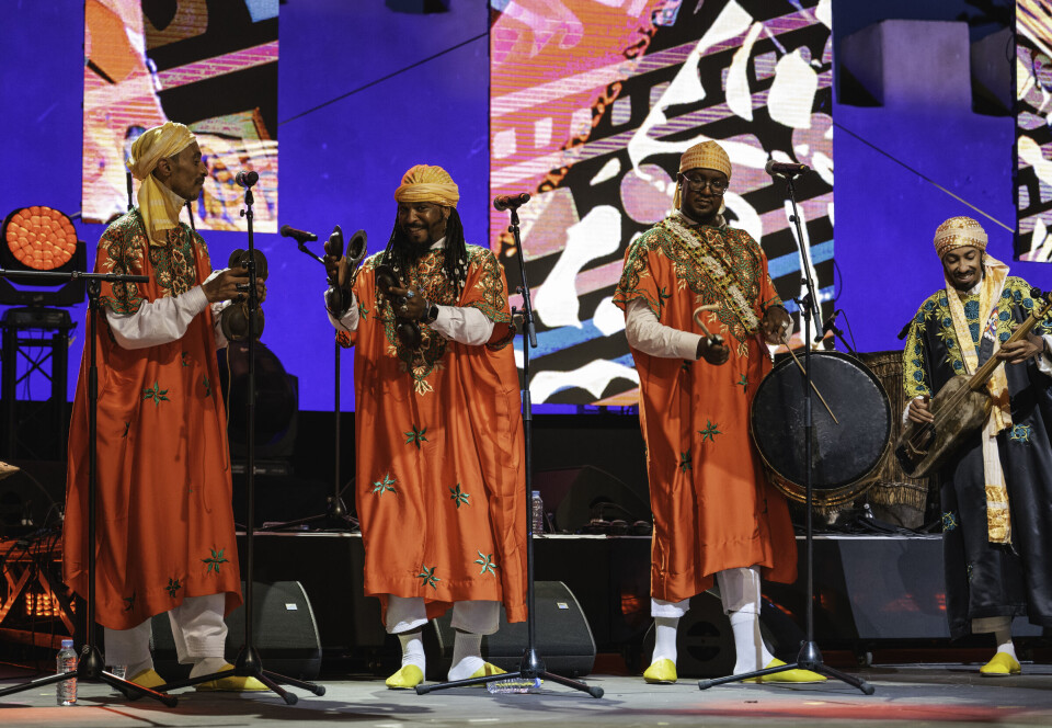 Four men are on stage in traditional red and green jellabas, two are playing the qraqeb, one the tleb drum, and one the guembri.