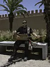 A man in his thirties, all dressed in black, is sitting outside on a stone bench, holding a guembri and smiling at the camera.