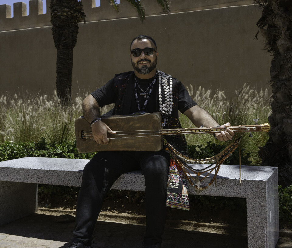 A man in his thirties, all dressed in black, is sitting outside on a stone bench, holding a guembri and smiling at the camera.