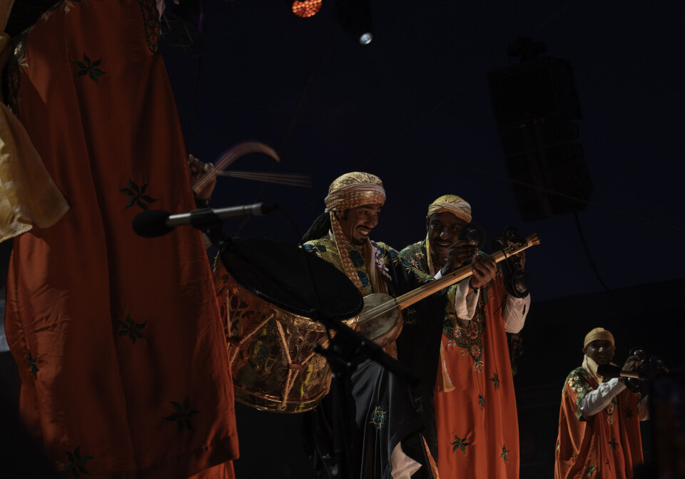 Four men are on stage, wearing traditional Moroccan garments in red and green. One is playing a gumebri, two the qraqeb, and another a big drum.