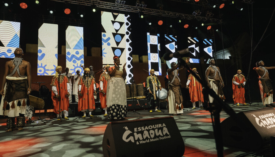 Several Moroccans and Rwandans are on stage together, wearing their respective traditional attire: the Moroccans are wearing colorful jellabas, while the Rwandans are in brown-and-white garments with the men’s torsos exposed.