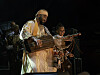 A man wearing white traditional Moroccan clothes is playing a guembri on stage, laughing with a boy wearing a brown traditional jellaba and dancing.