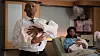 Jonasi stands in the foreground holding a swaddled newborn baby. Behind him, a woman sits in a hospital bed holding another baby.