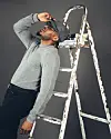 Alikiba poses beside a white ladder stacked with books, wearing a grey sweater, black trousers, glasses and a grey hat.