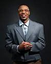 Alikiba smiles in a studio portrait, wearing gold-framed glasses, a blue-grey suit jacket, and a light blue shirt.