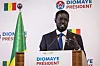 Bassirou Diomaye Faye speaks at a podium during a press conference in Dakar with Senegal flags and campaign branding behind him.
