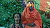 Hiba Elgizouli is sitting next to a Sudanese woman wearing an orange toub. Her eyes are covered with orange flowers, and her face is painted with turquoise makeup.