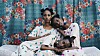 Three sisters in white dresses with green, blue, and purple dots sit in front of a blue curtain with red flowers, wearing lightbulb earrings and holding each other.