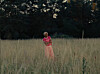 A woman in pink stands in tall grass with trees in the background.