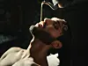 A young, shirtless North African man leans backward as tea is poured into his open mouth from a silver teapot.