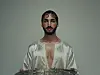 A man wearing a white caftan is holding a large silver tray. His hair is perfectly styled, and his lips are shiny with lipgloss.