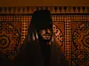 A man sitting in a red room in front of a wall of Moroccan tiles, his face half-hidden under a cloth and a tarboush, his lips shining with lipgloss in the dim light.