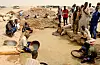 Men and boys pan for gold beside mounds of earth in a dusty Sudanese village.