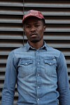 Man wearing a denim shirt and red striped cap standing before a corrugated metal background.