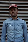 Man wearing a denim shirt and red striped cap standing before a corrugated metal background.