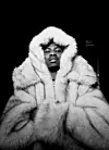 Black-and-white portrait of a person wearing a large fur coat with a hood against a dark background.