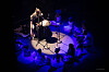 Person at a microphone surrounded by seated attendees in a circular blue-lit performance space.