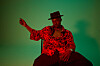 Ben September sits on a chair and gazes at the camera in a new promo image. He’s showered in red light, dressed in a red shirt, and dons a black hat.
