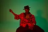 Ben September sits on a chair and gazes at the camera in a new promo image. He’s showered in red light, dressed in a red shirt, and dons a black hat.