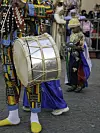 A man is carrying a large drum; behind him, a little boy is holding large qraqeb. Both are dressed in colorful traditional attire.