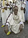 An elderly man dressed in cream-colored traditional Moroccan attire is squatting comfortably on the street, looking directly at the camera.