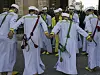 Nine men wearing light blue traditional Moroccan attire and daggers at their waists are forming a circle, holding hands and dancing.