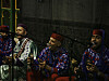 Four men are sitting on the floor and singing into microphones. Three of them are playing qraqeb, and one the guembri. The qraqeb players are wearing blue and red, and the guembri player is wearing green.