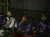 Four men are sitting on the floor and singing into microphones. Three of them are playing qraqeb, and one the guembri. The qraqeb players are wearing blue and red, and the guembri player is wearing green.