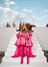 Salima Chica stands on white steps in a bright pink outfit, with blonde hair and a circular structure behind her.