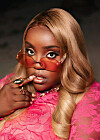 Close-up portrait of Salima Chica wearing red-tinted sunglasses, blonde hair, gold jewelry, and a bright pink lace outfit.