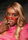 Close-up portrait of Salima Chica wearing red-tinted sunglasses, blonde hair, gold jewelry, and a bright pink lace outfit.