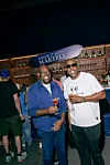 Two men stand smiling with cocktails in front of a Martell sign and bar shelves.