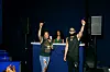 Two hosts stand on a stage with raised arms, with a Martell-branded backdrop and speakers behind them.