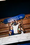 Three people pose at a Martell event with bottles on shelves and a large blue sign reading Make it with Martell.