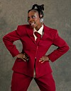 Woman in a red suit stands with hands on hips against a gray background.