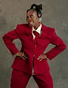 Woman in a red suit stands with hands on hips against a gray background.