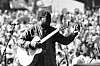 Black-and-white image of Vusi Mahlasela on guitar performing during an outdoor concert, with microphones in the foreground and a blurred audience behind.