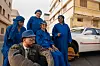 A man wearing a grey leather jacket and blue hat is sitting with four elderly women, all wearing jellabas and blue headscarves.